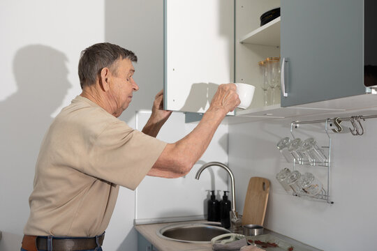 Senior man standing in modern kitchen pouring hot water from electric kettle into mug on table. - Powered by Adobe