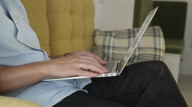 Expectant man in light blue shirt and denim shorts working on laptop computer seated on mustard yellow couch with plaid pillow. Remote work and pregnancy lifestyle at home.