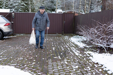 Elderly man walking with cane across brick courtyard, carrying shopping bag, winter setting, residential gate background.
