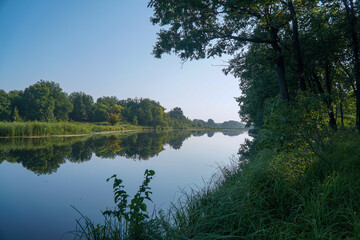 A small river or lake with blue water and a green forest on the shore under a blue sky. Summer landscape