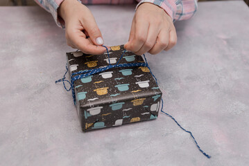 Hands of a young woman tying a blue string around a gift box. The box is decorated with colorful kitchen utensils. A light gray surface is visible.