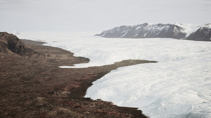 A breathtaking scene unfolds as a massive glacier stretches across the land, contrasting with the rocky terrain. The soft light of dawn illuminates the icy surface and nearby mountains.