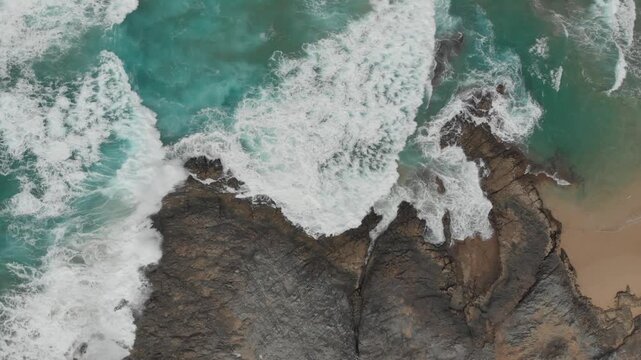 Aerial view of the powerful ocean waves crashing against rugged coastal rocks, with clear turquoise water meeting the sandy beach, creating a serene yet dramatic landscape.