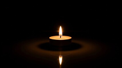 A single, warm, glowing candle flame flickers gently, casting a soft orange light and reflections in the darkness.