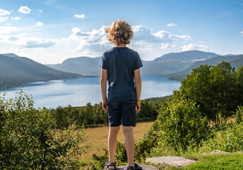 A boy looks out over Gjevillvatnet lake surrounded by mountains and green hills. The scene shows a bright summer day with blue sky and soft clouds, in Oppdal, Tr&oslash;ndelag Norway