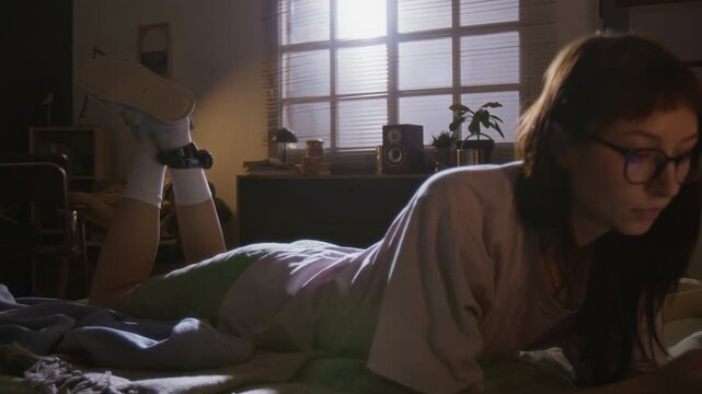 Young woman in glasses, wearing house arrest ankle bracelet, lying on her stomach in bed and using smartphone