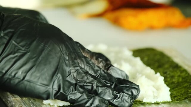 Chef hands in latex gloves spreads boiled rice on nori sheet at table closeup. Japanese cuisine professional prepares sushi in kitchen