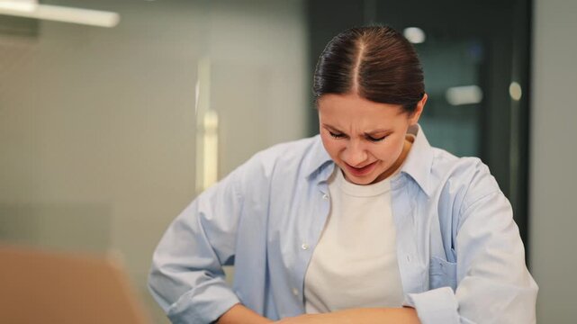 Young woman sitting in office suffering from severe abdominal pain. Feeling unwell, discomfort, and distress due to sudden illness or menstrual cramps. Seeking relief from persistent stomach ache.