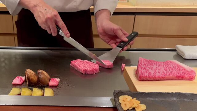 Teppanyaki chef trimming rich fat from Kobe A5 beef on hot iron griddle with arranged mushrooms and sliced vegetables, capturing precise start of cooking process