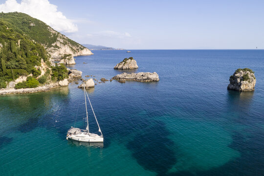Aerial view of a white sailboat gliding through the turquoise waters near rocky outcrops and verdant hillsides, Panagia, Thasos Regional Unit, Greece.