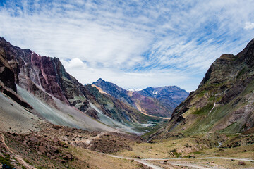 View of rugged mountains rise in a symphony of ochre, brown, and grey hues beneath a vast sky streaked with wisps of white clouds, San José de Maipo, Santiago Metropolitan Region, Chile.