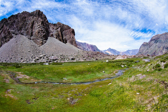 View of a vibrant green meadow bisected by a winding stream, nestled beneath towering rocky mountains against a bright, patterned sky, San Jos&eacute; de Maipo, Santiago Metropolitan Region, Chile.