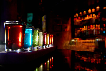 A row of nine vibrant, glowing mixed shots on a dark bar counter. Captures the high-contrast...