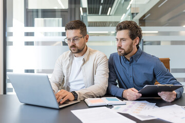 Two business colleagues working together, analyzing data on a laptop and reviewing documents, demonstrating teamwork and professional collaboration in a modern office environment
