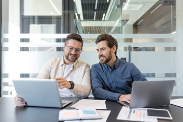 Two professional businessmen collaborating and discussing work using a laptop in a modern office meeting room, focusing on data and reports for successful teamwork
