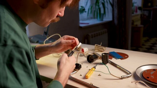 Skillful upcycling master working at home workshop on renewing vintage lamp, uses screwdriver and pliers to repair old light bulb socket, crimps and tightens wire contacts, prepares new wiring.