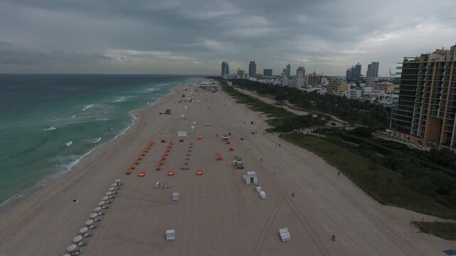 Aerial view of a sandy beach meeting the turquoise ocean under a moody sky, with rows of umbrellas and the cityscape in the background, Miami Beach, Florida, United States.