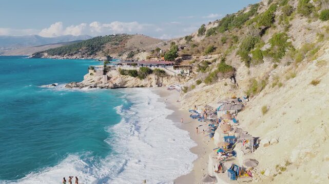 Aerial view of the beach with turquoise waters meeting the shore, a resort with tan structures and sunbathers, along a rocky coastline, Ksamil, Qarku i Vlores, Albania.