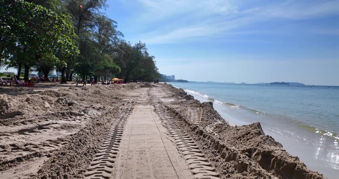 Shoreline construction scene featuring heavy tread marks and distant excavator against a calm blue sea.