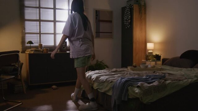 Young woman in glasses, wearing house arrest ankle bracelet, getting out of bed and looking out of window after waking up