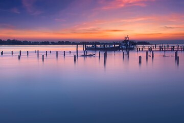 Serene sunset over calm water with silhouetted wooden pilings and moored boats, reflecting vibrant pink and orange sky colors in a long exposure for silky smooth surface.
