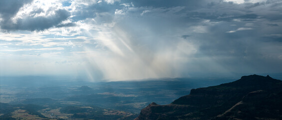 Aerial view of sun rays piercing through dark clouds over the undulating landscape, casting shadows on the hills, Nashik, Maharashtra, India.