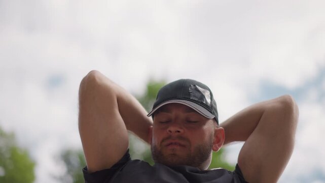 man in cap relaxing outdoors arms behind head male golfer taking restful pause after swing sky trees background warm sunlight casual polo beard contemplative expression park setting summer mood candid