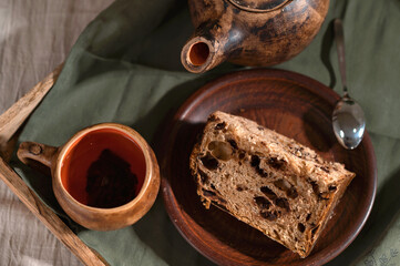 Piece of panettone sweet cake on plate, brown clay cup of tea and teapot, green linen napkin. Delicious traditional holiday bakery with raisins