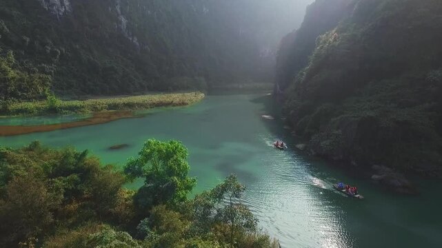 A drone flies over the Trang an Valley, a large body of water, with mountains covered in dense forest to the right and left, and boats with people can be seen in the water.