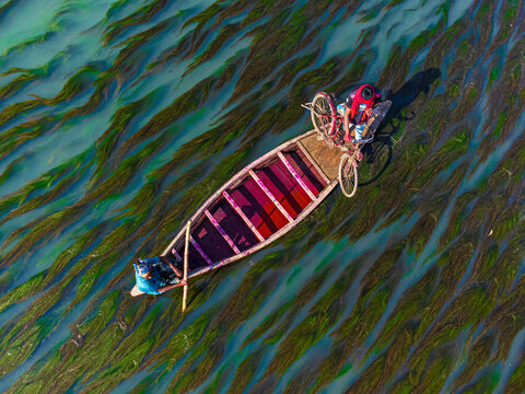 Rajshahi, Bangladesh - 13 December 2025: Aerial view of a vibrant boat cutting through the murky green waters, carrying a bicycle and passengers under the watchful eye of the sun.