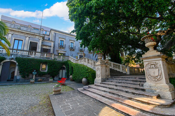 CATANIA, ITALY. The lush courtyard of Villa Cerami, a historic 18th-century palace currently housing the Faculty of Law, featuring a large palm tree, stone statues, and a grand stairc