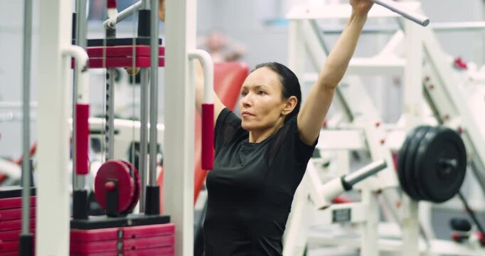 Caucasian woman doing exercise on a lat machine in the gym. Pulldown reps during gym workout session. Middle-aged woman works her back with controlled lat pulldown reps in the gym.
