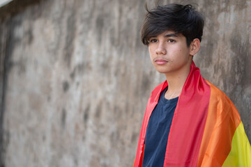 A young person wrapped in a rainbow flag, symbolizing LGBTQ+ pride, stands against a textured wall. The rainbow flag represents diversity and the spectrum of human sexuality and gender.