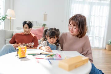 Mother helps child with art project while father plays guitar in living room at home during afternoon