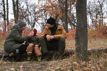 Fototapeta premium A family of three: a mother, father, and son on a picnic in nature in cold weather, drinking tea from a thermos and eating together, relaxing in the fresh air in the forest