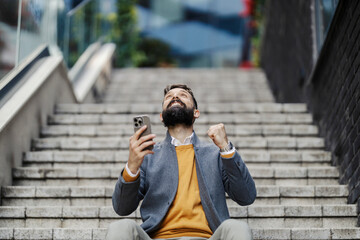 Cheerful successful businessman sitting on staircase with cellphone in hands and celebrating victory.