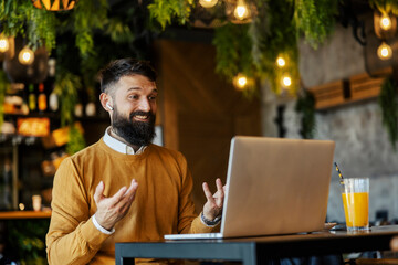 Portrait of friendly cheerful businessman sitting in cafeteria and having conference call with colleagues on a laptop.