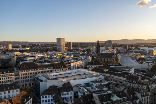Aerial view of the cityscape with a blend of modern and traditional buildings under a warm sky, highlighting the steeple of a church, Basel, Basel City, Switzerland.