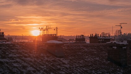 Obraz premium Bathing snow-dusted sloped tiled rooftop at sunset, sun near horizon showing chimneys and masts