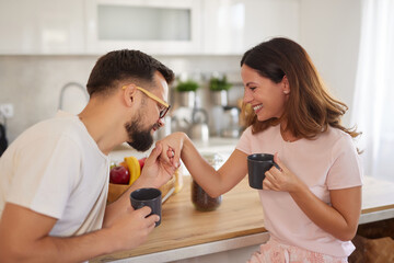 A couple begins their day together in a bright kitchen, laughing and sipping coffee. They share a light moment, showing affection and joy in a relaxed atmosphere, surrounded by fruit and daylight.