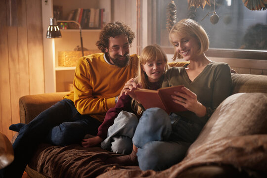 A joyful family sits closely on a sofa, reading a storybook together. The warm glow of a lamp enhances the cozy atmosphere of their living room in the evening.