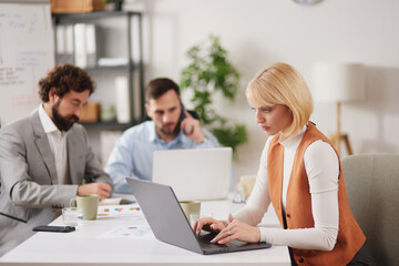 Obraz premium Individuals work together in an office environment. One person types on a laptop while others discuss ideas on phones and papers. The scene shows collaboration in real time.