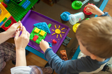 A child and an adult are sitting at a table, using play dough to create a house and sun on a purple tray. Other play dough containers and some toys are nearby on the table.