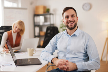 A group of professionals is engaged in a meeting at a bright office setting. One man is smiling while seated at a table with a laptop. A woman is focused on taking notes.