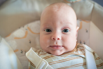 A cute infant, wearing a striped shirt and strapped into a white bouncer, gazes directly at the camera. The baby has light skin, round cheeks, and bright blue eyes.
