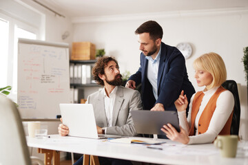 A business team is involved in a brainstorming session. Three team members share ideas at a table in a modern office. Laptops and tablets are in use for collaboration.