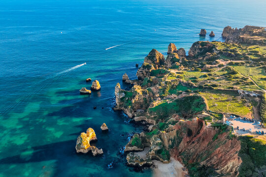Aerial view of dramatic cliffs meeting the turquoise sea, boats cutting through the water near Praia do Camilo, Lagos, Faro, Algarve, Portugal.