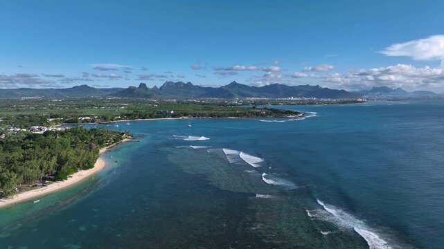 Drone shot in Mauritius from Pamplemousses facing Baie-du-Tombeau, with Port Louis visible in the far distance. Reef breaks trace the shoreline as mountains rise across the calm tropical backdrop.