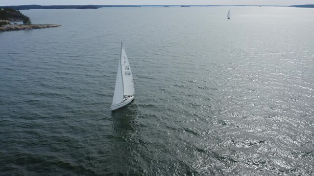 Sailboat navigating with full sails close to shore in Finland South West archipelago. Small islands in the background. Aerial drone view circling around.