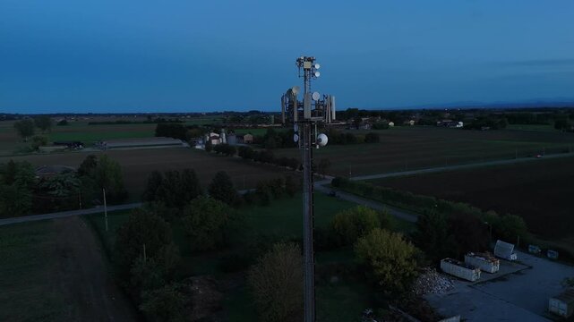Drone orbit around a cell tower in the Po Valley countryside, northern Italy, revealing surrounding fields, rural roads and a nearby waste‑management yard at dusk.